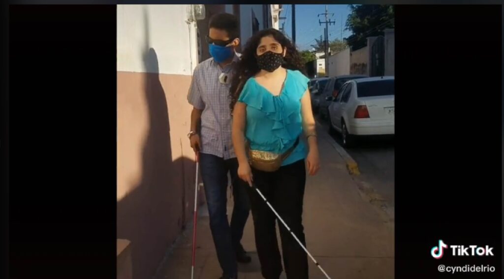En la imagen se ve la captura de un video en donde en la que se muestran  dos personas jóvenes con discapacidad visual caminando por la calle.