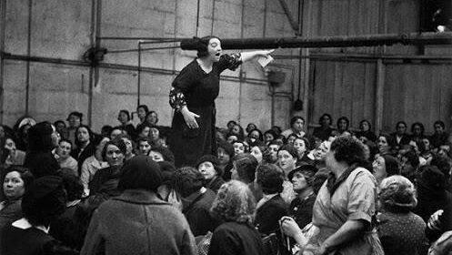 Imagen de una trabajadora encima de una mesa dando un discurso a las otras trabajadoras de la fabrica. 
